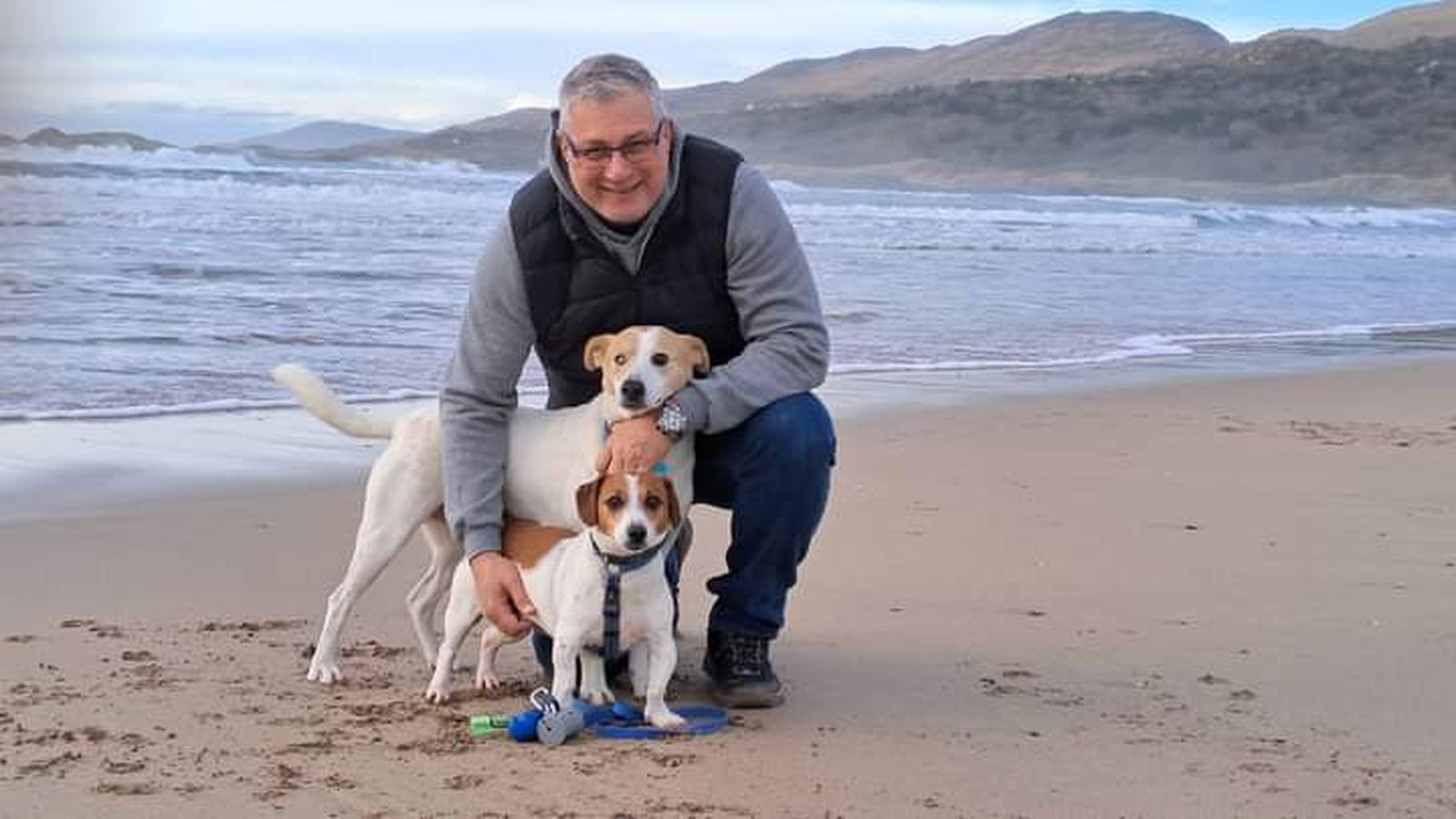 Albert crouching on a Kerry beach with his two Jack Russells, mountains and surf behind